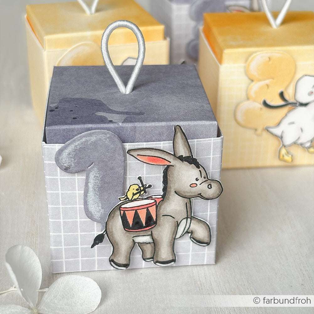 Alexandra Renke Geschenkbox mit einem Cartoon-Esel, gelbem Vogel, lila Schwanz und Trommel. Hergestellt aus hochwertigen Materialien mit weißer Oberschleife und detailreichen Motiven für zusätzlichen Charme.