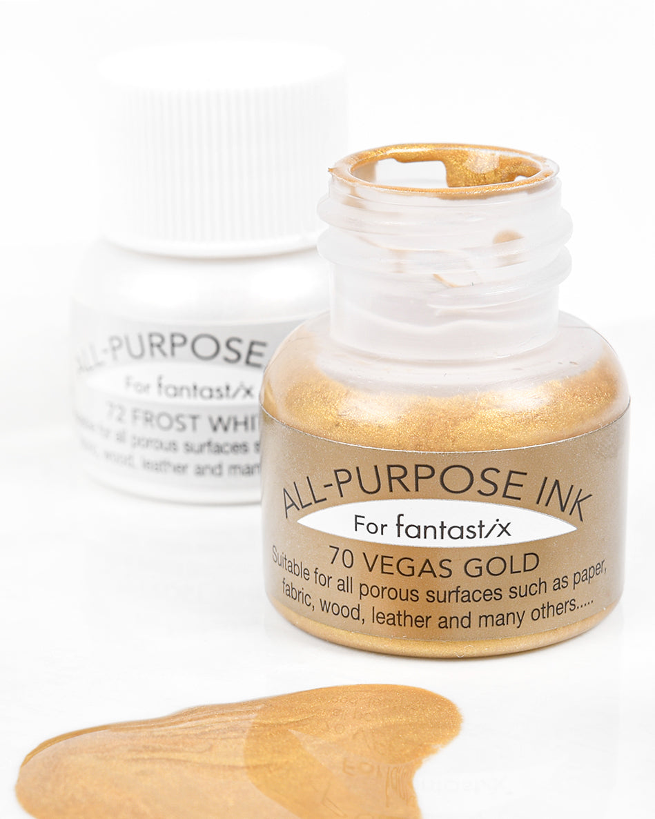 Geöffnetes Gefäß mit goldener Allzwecktinte, daneben verschüttete goldene Tinte; weißes Tintenfass im Hintergrund unscharf.