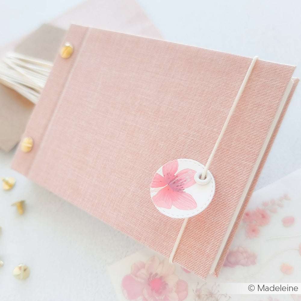 Ein rosafarbenes, stoffbezogenes Tagebuch von ☆ mit einem geblümten Anhänger, einem elastischen Band und goldenen Brads. Hergestellt aus nachhaltigem Buchleinen, vereint es Stil und Umweltbewusstsein in einem eleganten Accessoire.