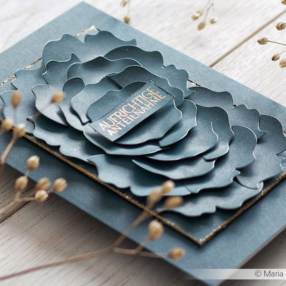 Eine mehrschichtige blaue Papierblumen-Grußkarte von Alexandra Renke mit deutschem Text und Photopolymer-Stempel-Akzenten wird auf einer Holzfläche mit getrockneten Blumen in der Nähe ausgestellt.