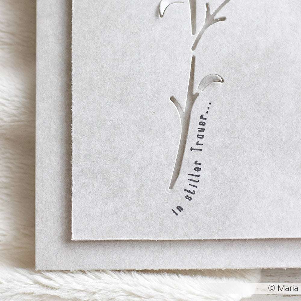 Nahaufnahme einer grauen Karte mit einem ausgeschnittenen Blumenmotiv, hergestellt mit Alexandra Renke Photopolymer Stempel, auf dem der Text "in stiller Trauer..." aufgedruckt ist.