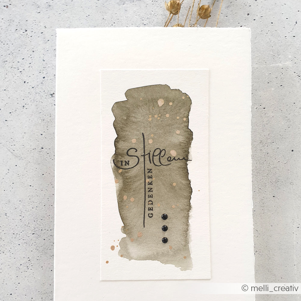 Eine Aquarellkarte von Alexandra Renke mit dem deutschen Text "In stillem Gedenken" auf grauem Hintergrund mit Farbspritzern, erstellt mit einem Clear Stamp Set für gestochen scharfe, elegante Schriftzüge.