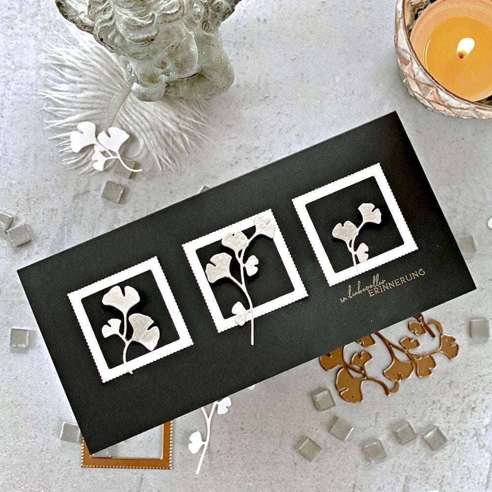 Schwarze Karte mit drei umrahmten weißen Ginkgoblättern, die mit einem Photopolymer-Stempel von Alexandra Renke hergestellt wurden, neben einer Kerze und Dekor auf einer grauen Fläche.
