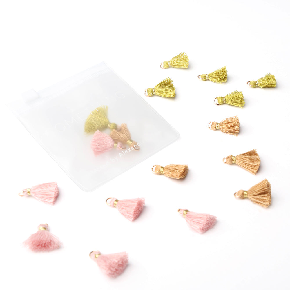 Kleine Quasten in Rosa, Gold und Grün von Alexandra Renke sind verstreut angeordnet, einige davon befinden sich in einem mattierten Plastikbeutel – ein bezauberndes Schmuckstück, das sich perfekt zur Verzierung von Buchbindearbeiten oder kreativen Projekten eignet.