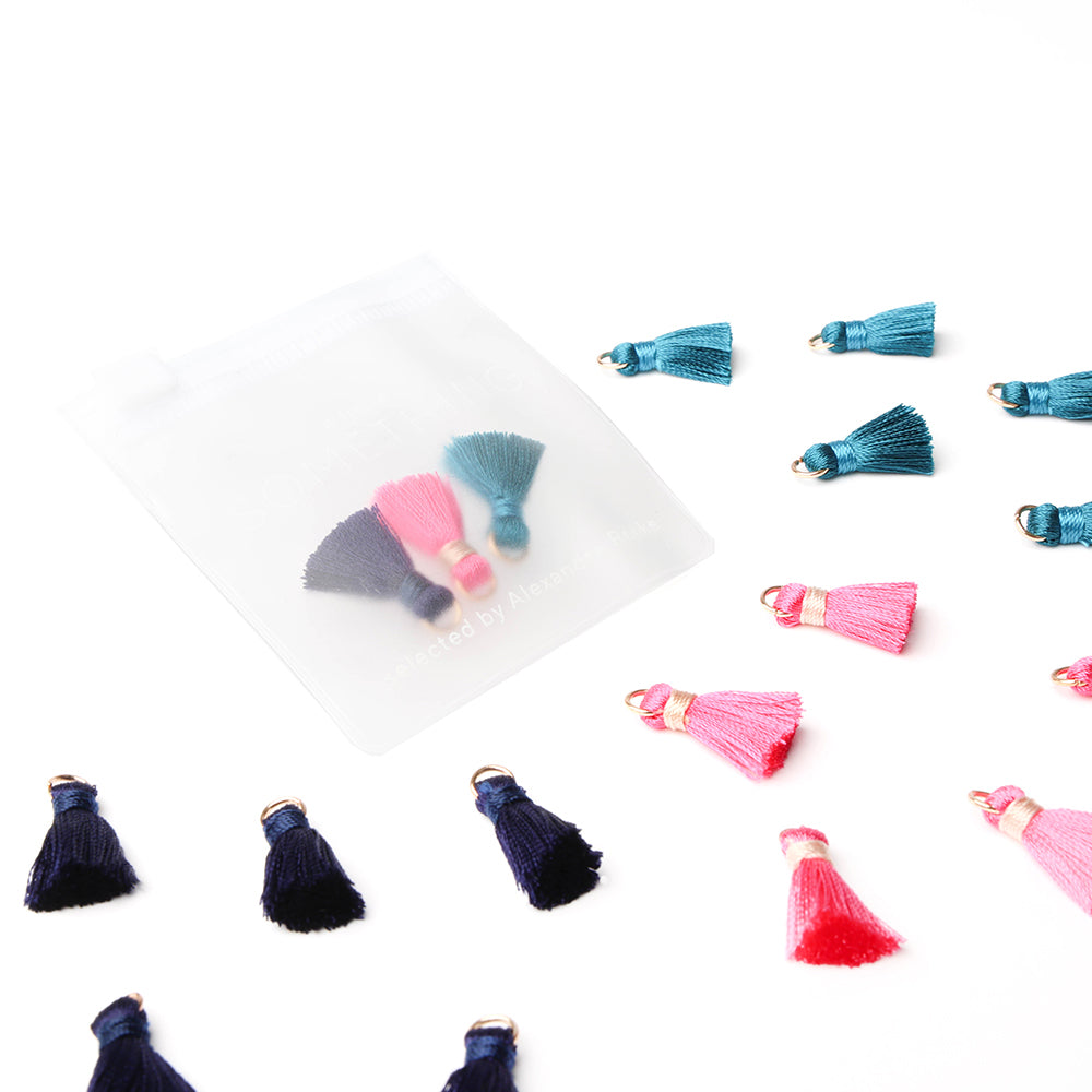 Die „Mini Tasseln Mix“ in Blau, Rosa und Marineblau von Alexandra Renke sind auf einem weißen Untergrund verstreut – einige davon befinden sich als charmantes Schmuckstück in einem kleinen durchsichtigen Beutel. Perfekt für Buchbindearbeiten und kreative Projekte!.