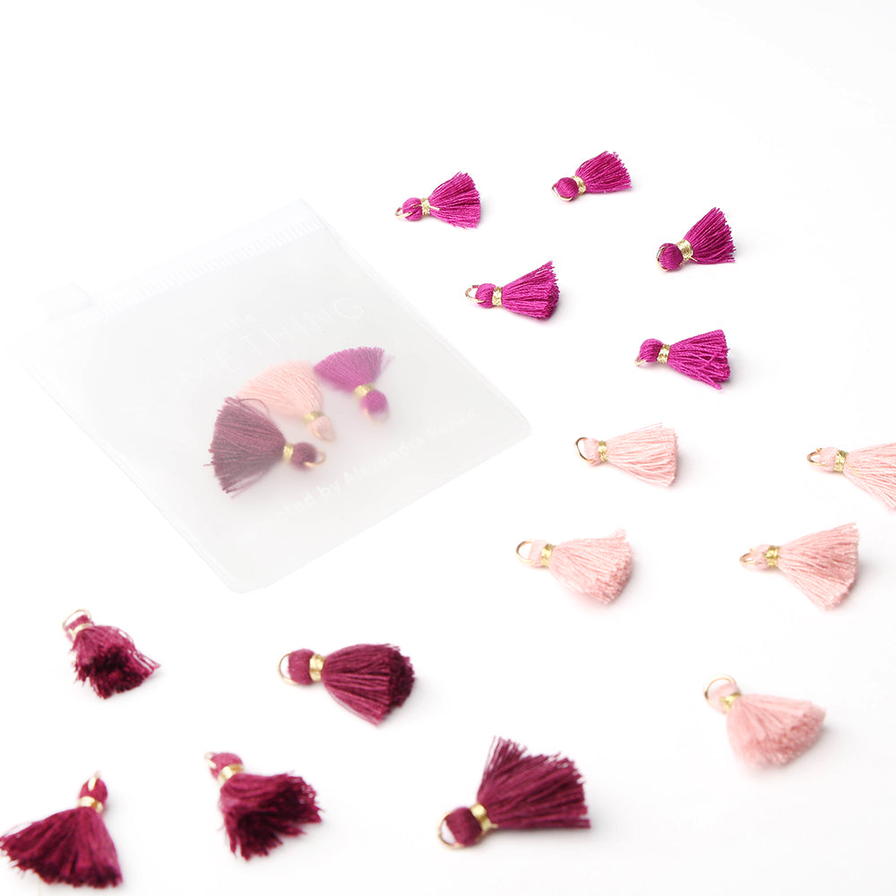 Kleine rosa und burgunderrote Quasten von Alexandra Renke sind auf einem weißen Untergrund verteilt, einige davon befinden sich in einem durchsichtigen Beutel – ein idealer Quasten-Mix für Schmuckstücke oder Buchbindearbeiten.