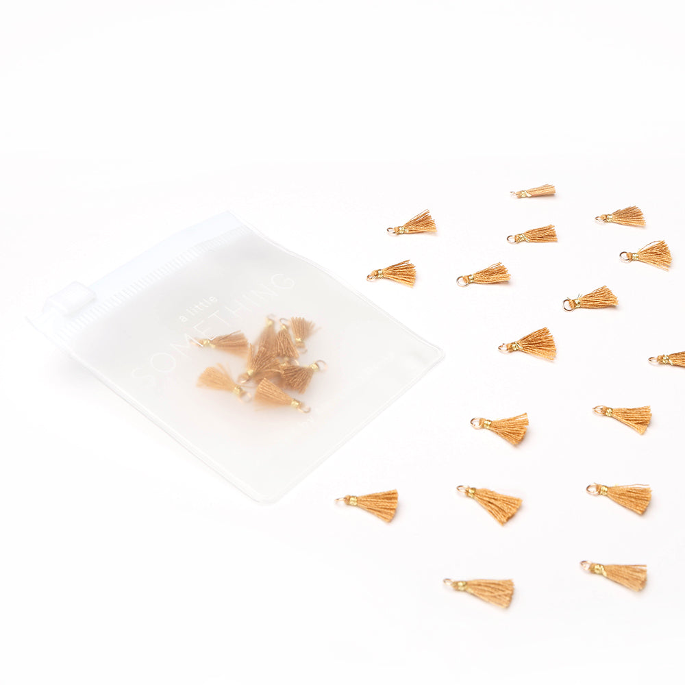 Kleine goldene Quastenanhänger und Tasseln Rehbraun von Alexandra Renke sind um einen transparenten Beutel auf weißem Hintergrund angeordnet - ideal für Buchbindearbeiten oder Schmuck selber machen.