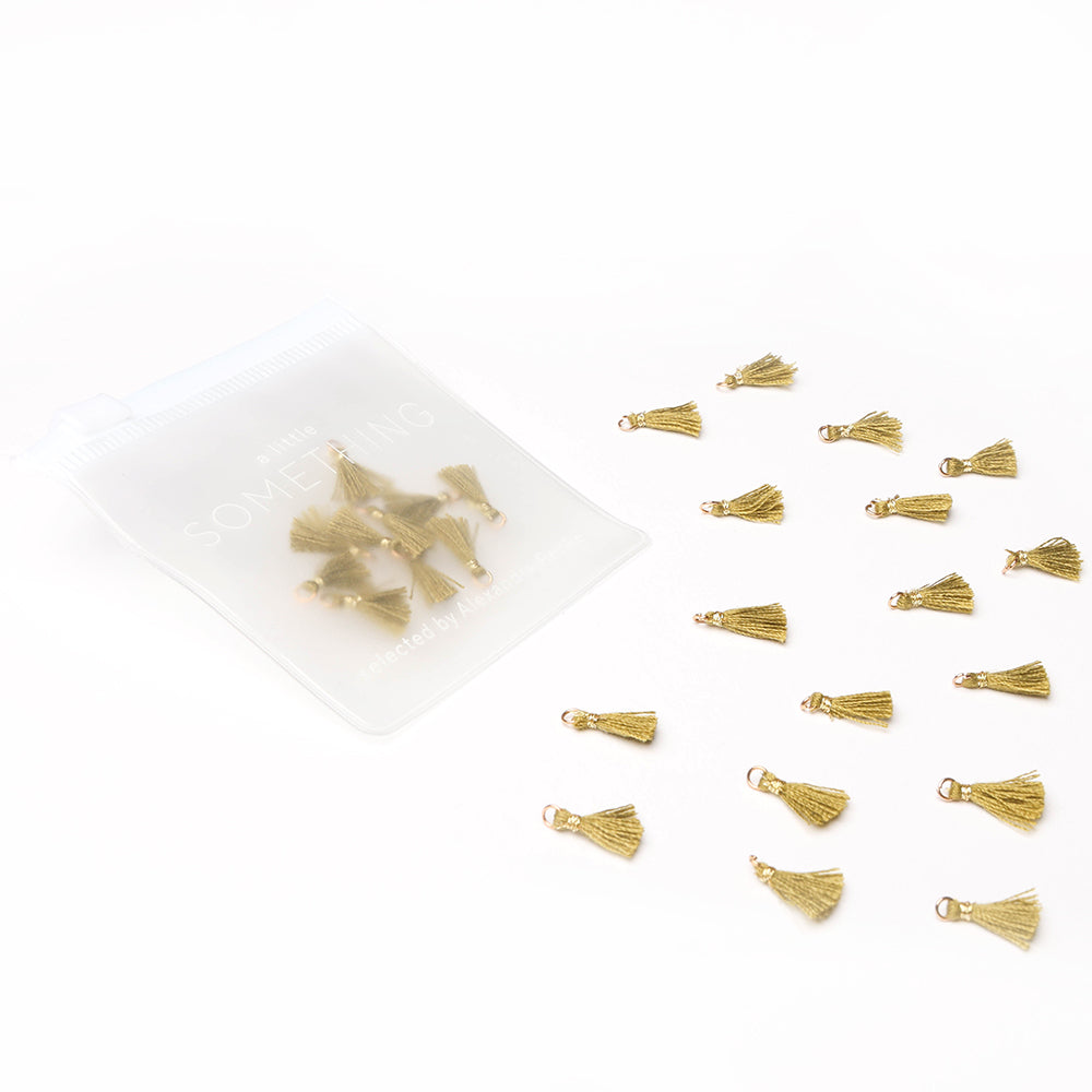 Die kleinen Tasseln Goldbraun von Alexandra Renke sind auf einer weißen Fläche verstreut, weitere befinden sich in einem durchsichtigen Beutel auf der linken Seite - ideal für kreative Papierprojekte und Buchbinderei.