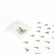 Die kleinen grünen Quastenanhänger von Alexandra Renke, die sich ideal für Papierprojekte und Buchbindearbeiten eignen, werden auf einer weißen Fläche ausgestellt, wobei mehrere von ihnen in einem gefrosteten Plastikbeutel stecken.