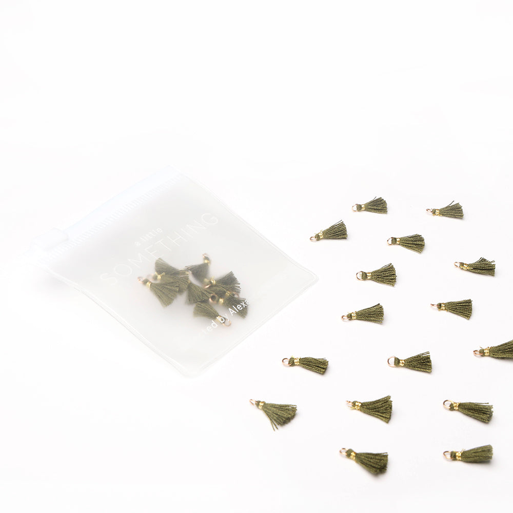 Kleine olivgrüne Tasseln von Alexandra Renke, verstreut neben einem durchsichtigen Reißverschlussbeutel mit weiteren Tasseln - perfekt für Buchbindearbeiten oder um jedem Schmuckstück eine einzigartige Note zu verleihen.