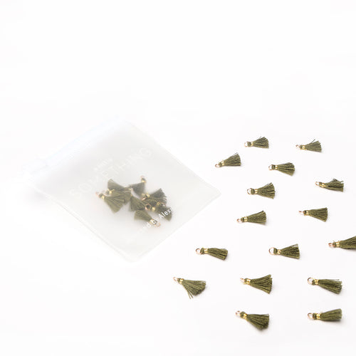 Kleine olivgrüne Tasseln von Alexandra Renke, verstreut neben einem durchsichtigen Reißverschlussbeutel mit weiteren Tasseln - perfekt für Buchbindearbeiten oder um jedem Schmuckstück eine einzigartige Note zu verleihen.