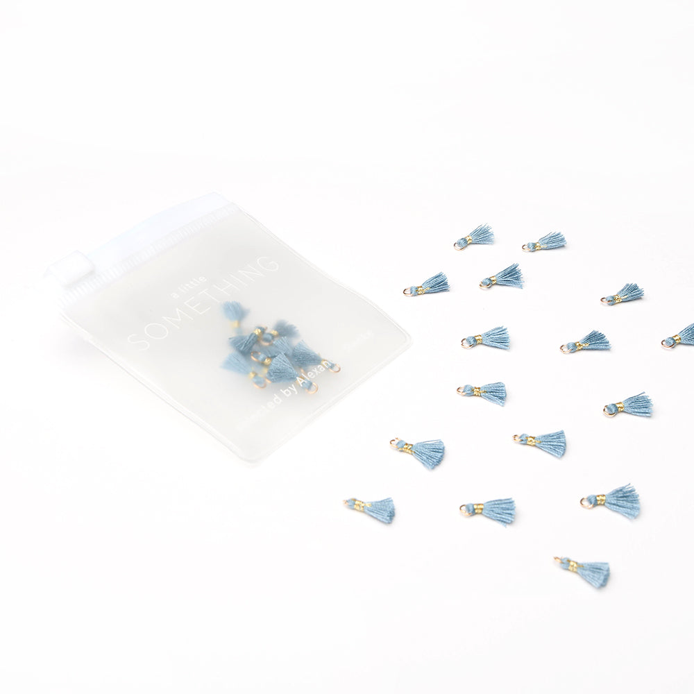 Die kleinen blauen Quastenanhänger von Alexandra Renke, die sich perfekt für Buchbindearbeiten oder als einzigartige Schmuckstücke eignen, werden neben einem durchsichtigen Beutel auf weißem Hintergrund präsentiert.