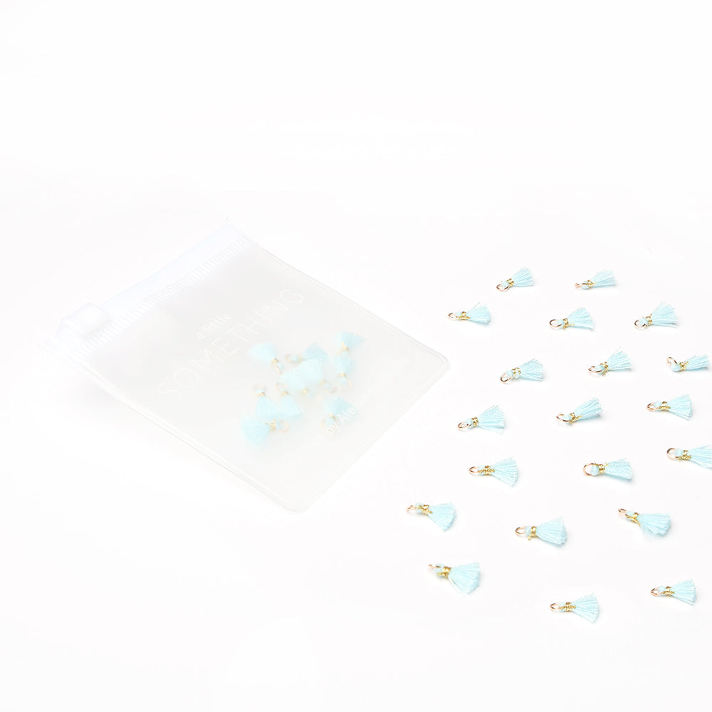 Kleine blaue Quastenanhänger von Alexandra Renke sind verstreut, mit mehreren in einem durchsichtigen Reißverschlussbeutel auf weißem Hintergrund - ideal für Buchbindearbeiten oder als einzigartiges Schmuckstück.