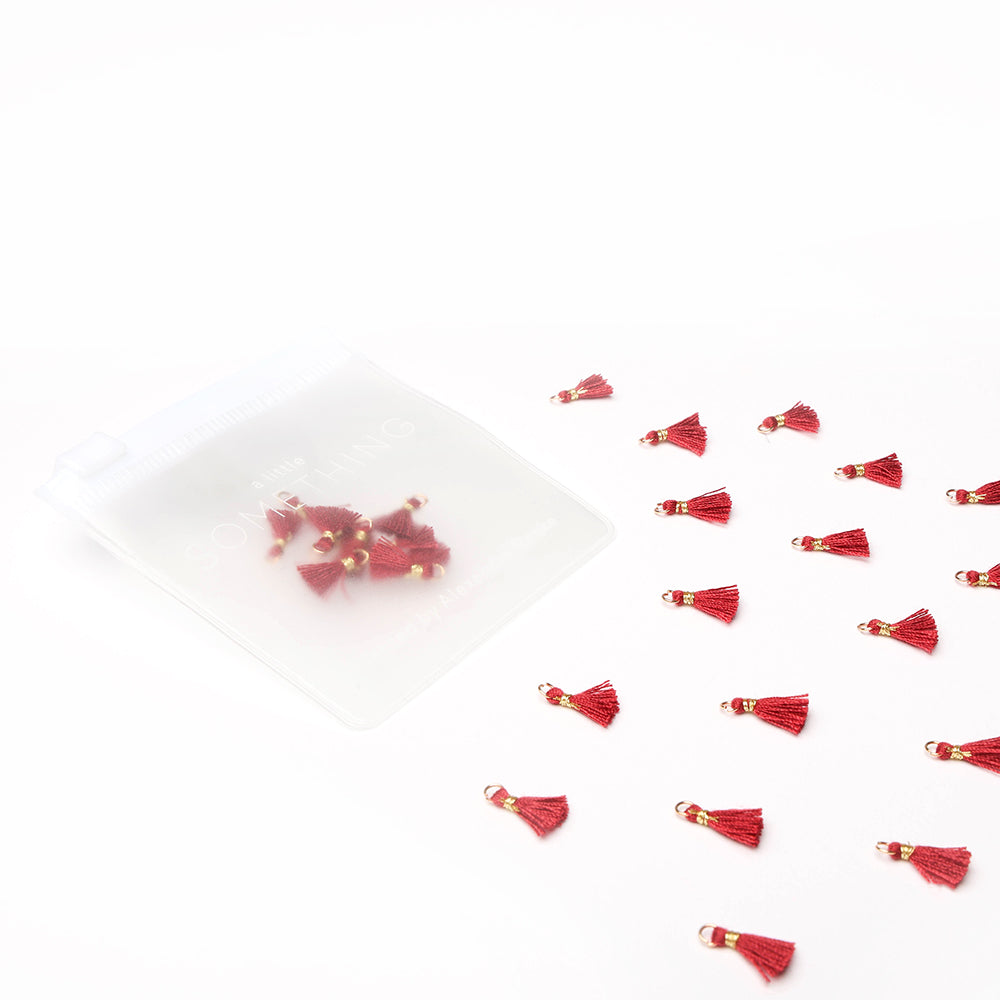 Die roten Tasseln Karminrot von Alexandra Renke sind neben einem durchscheinenden weißen Beutel auf weißem Hintergrund verstreut, ideal für Schmuck DIY oder Buchbinden Projekte.