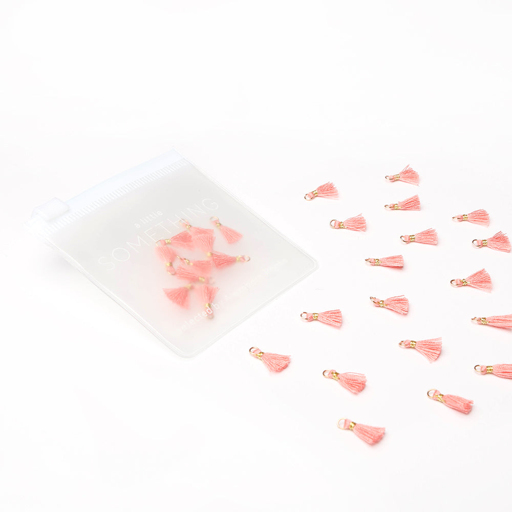 Kleine rosafarbene Quasten von Alexandra Renke, verstreut neben einer transparenten Reißverschlusstasche auf weißem Hintergrund - perfekt als Schmuckstücke oder für Buchbindungsprojekte.