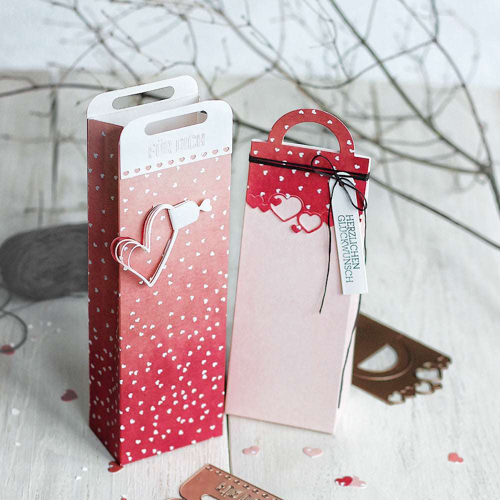 Zwei hohe Geschenkboxen von Alexandra Renke in Rot und Rosa mit Herzmotiven und Anhängern, hergestellt mit Dies und Motivstanze, ausgestellt auf einer hellen Holzoberfläche.