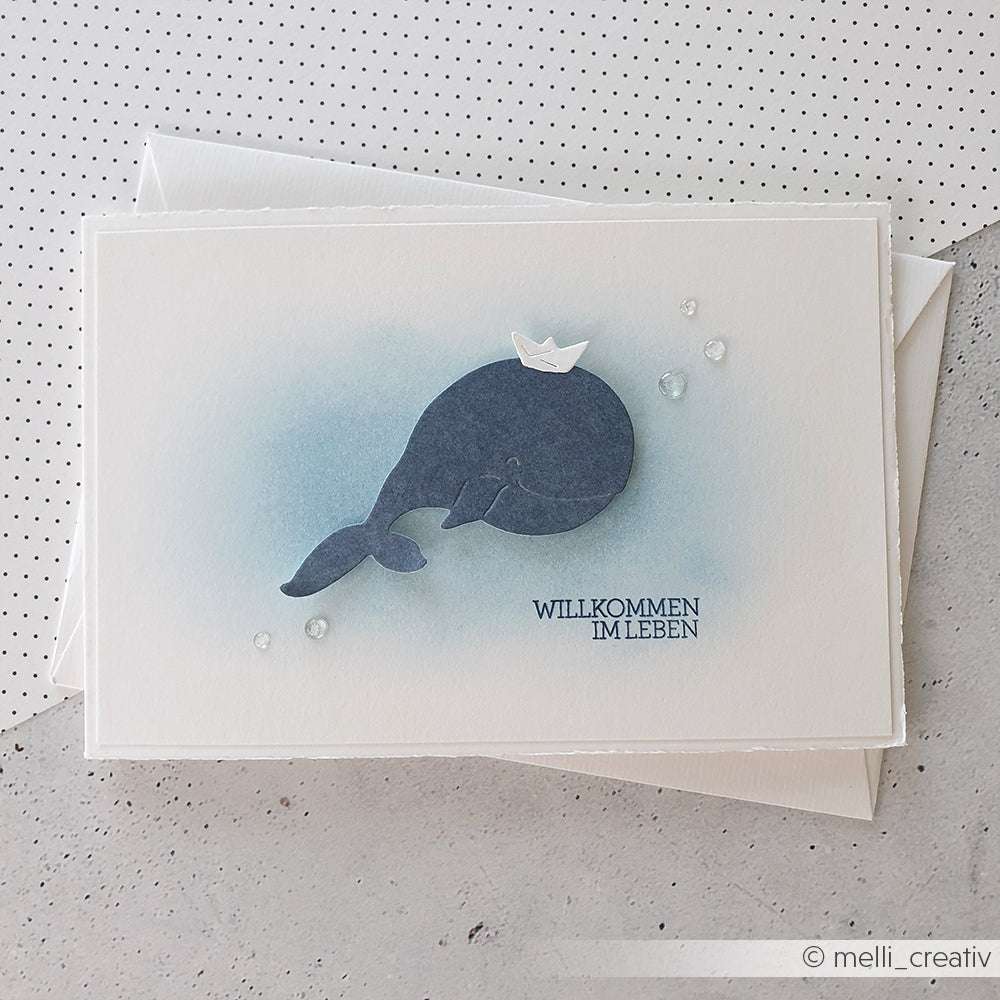 Grußkarte von Alexandra Renke mit einem blauen Wal in einem Papierhut, Wassertropfen und dem Text "WILLKOMMEN IM LEBEN" - eine perfekte Art, die besten Wünsche auf Deutsch zu übermitteln. Ideal für Fans von Clear Stamp Set oder Photopolymer Stempel.