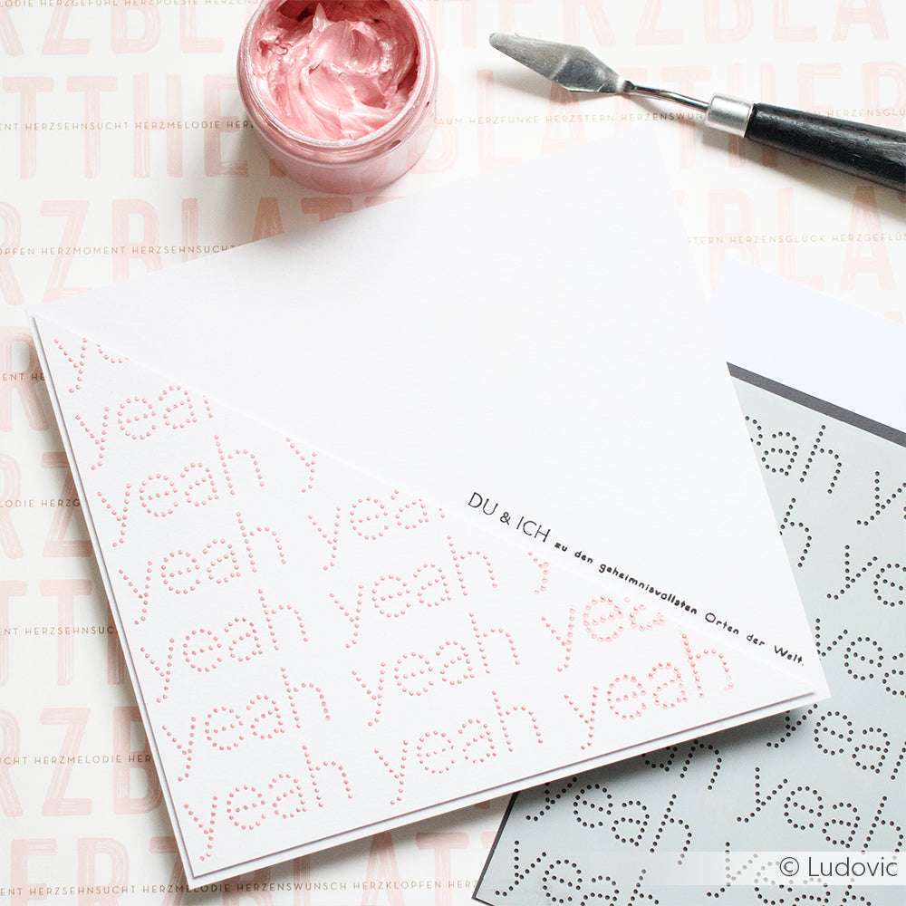 Eine weiße Karte von Alexandra Renke mit "yeah yeah yeah" in rosafarbenen Punkten und deutschem Text, hergestellt mit Schablonen, steht neben einem Glas mit rosafarbener Farbe und Kunstwerkzeugen.