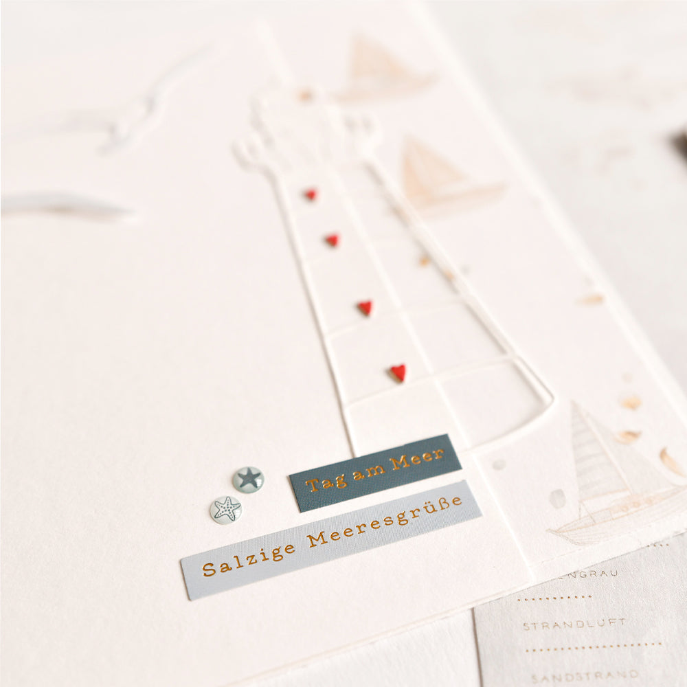 Nahaufnahme einer weißen Karte von Alexandra Renke mit geprägten Motiven eines Leuchtturms und einer Möwe sowie den Texten „Tag am Meer“ und „Salzige Meeresgrüße“ – ein bezauberndes Design für alle, die die wohltuende Meeresbrise lieben.