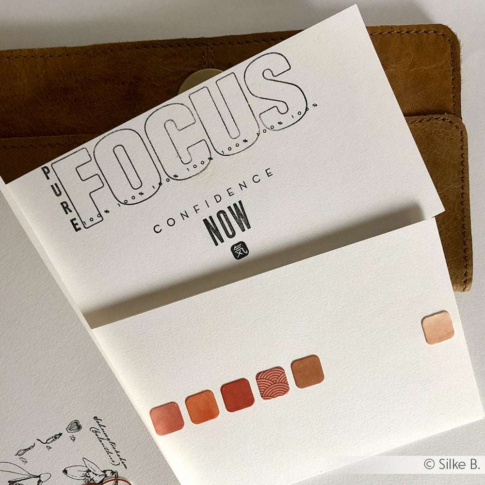 Ein aufgeschlagenes Notizbuch mit der Aufschrift PURE FOCUS CONFIDENCE NOW und roten quadratischen Aufklebern darunter eignet sich perfekt für die Kombination mit Ihrem Lieblings-Photopolymer-Stempel von Alexandra Renke oder einem klaren Stempelset auf einem Acrylblock.