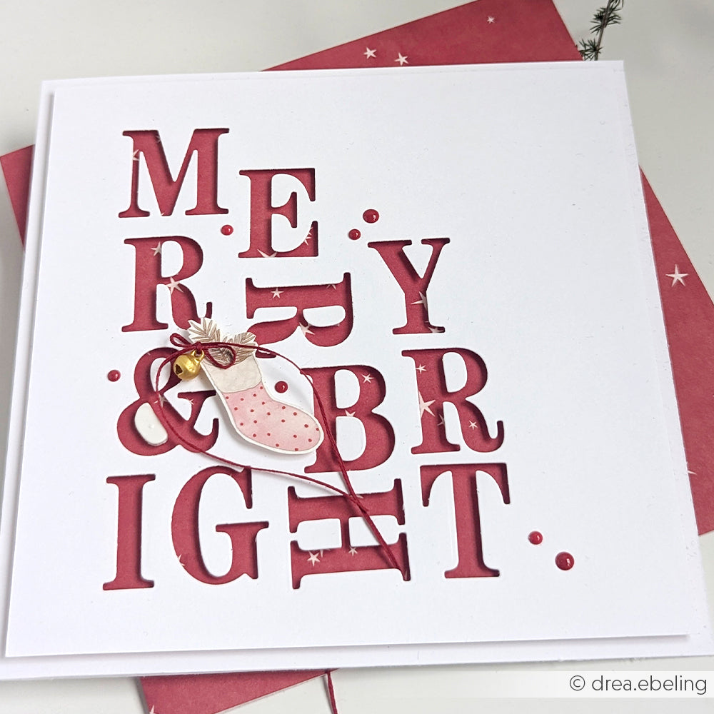 Weiße Karte mit ausgeschnittenem Text MERRY & BRIGHT, hergestellt mit Alexandra Renke Serifen Alphabet Stanzsysteme, und einer rosa Weihnachtsstrumpfdekoration auf rotem Hintergrund.