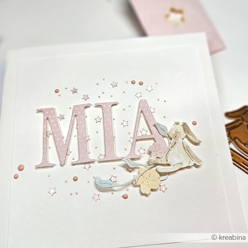Eine Karte mit MIA in rosafarbenen Buchstaben, hergestellt mit Alexandra Renkes Motivstanze Serifen Alphabet, verziert mit einem Häschen, Blumen und kleinen Sternen auf weißem Hintergrund.