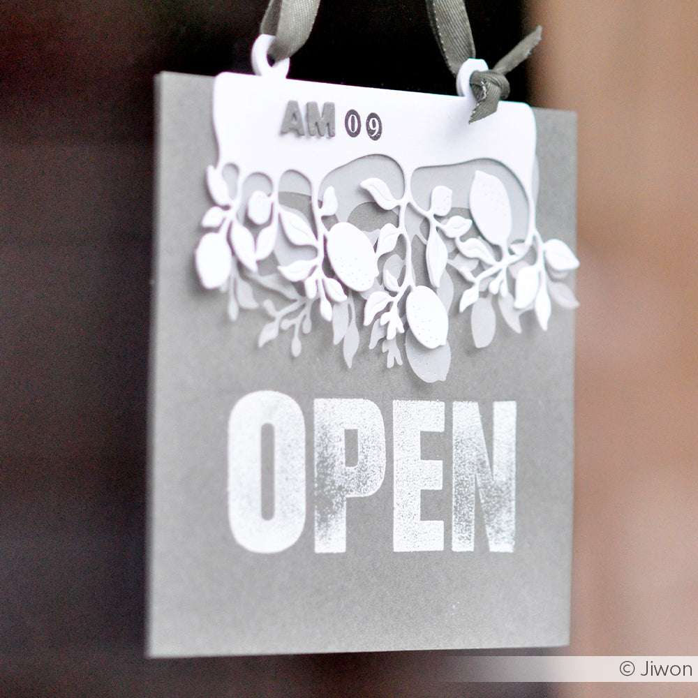 Graues OPEN-Schild mit weißen Zierblättern und dem Text "AM 09", das an einem dunklen Band hängt - im Stil der Block-Alphabet Groß Klarsichtstempel von Alexandra Renke für einen klaren, modernen Look.