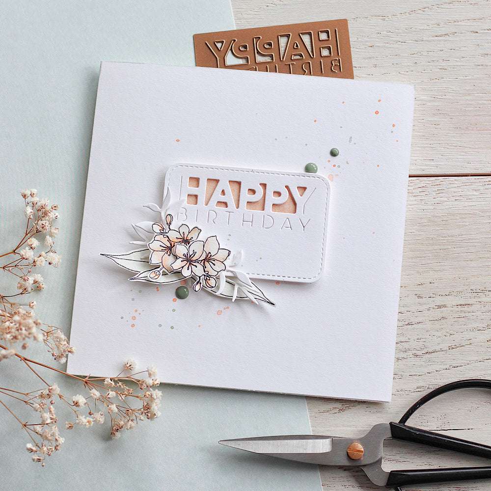 Quadratische Geburtstagskarte von Alexandra Renke mit HAPPY BIRTHDAY, floralem Design auf Designpapieren, Trockenblumen, Schere und einer Motivstanze abgerundetes Kärtchen auf hellem Untergrund.