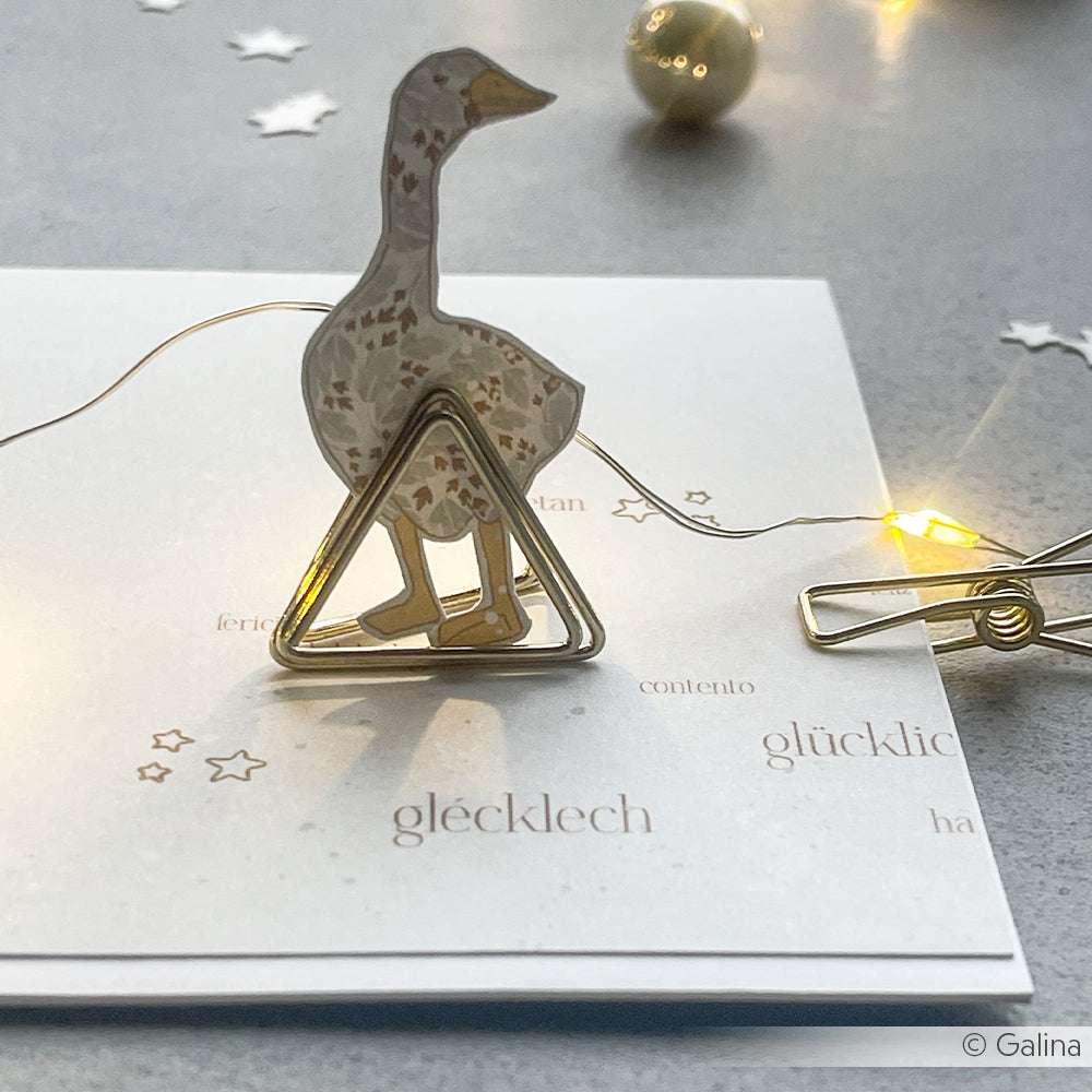 Eine Papiergans steht in einer dreieckigen Büroklammer auf einer Karte mit Lichtern, Sternenkonfetti und Alexandra Renke Clear Stamps wie Sternenbaumler in der Nähe.