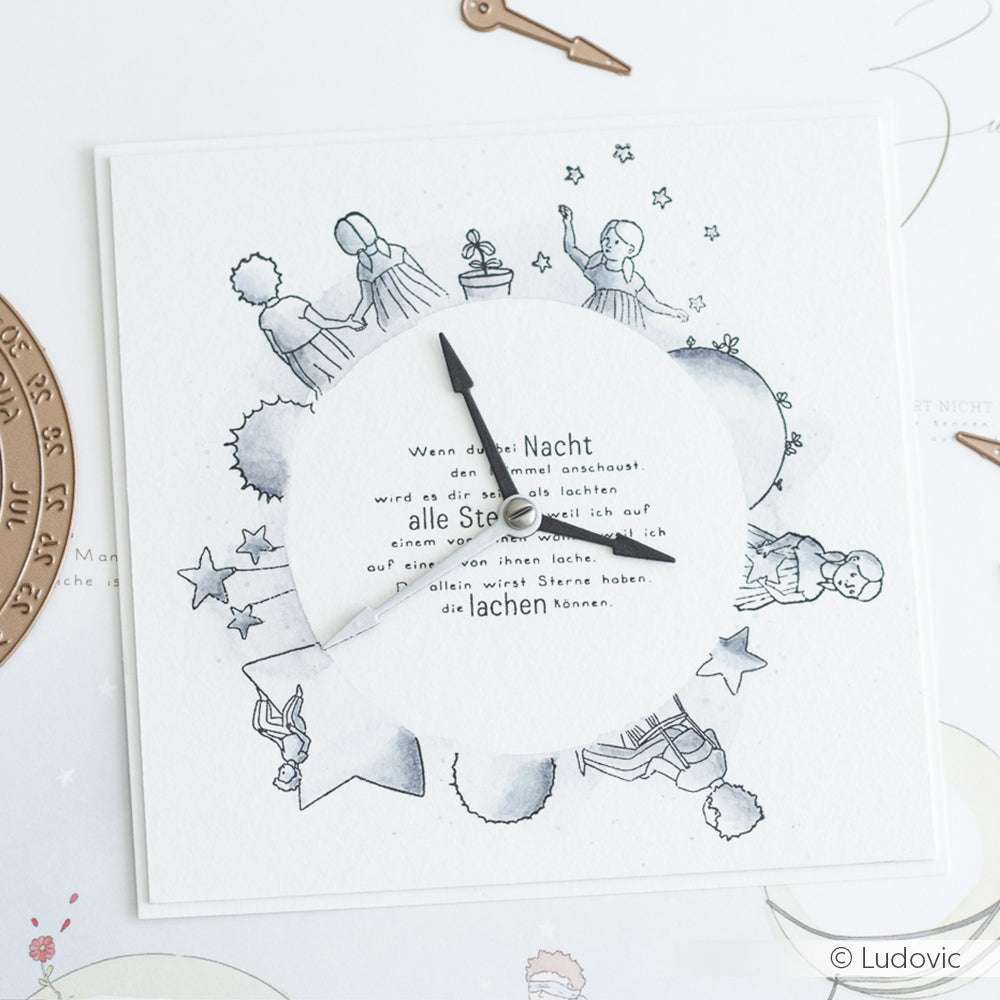Eine handgefertigte Uhr von Alexandra Renke mit Illustrationen des Kleinen Prinzen und deutschem Text in der Mitte, hergestellt mit Photopolymer-Stempel für gestochen scharfe Details auf einem weißen Hintergrund.
