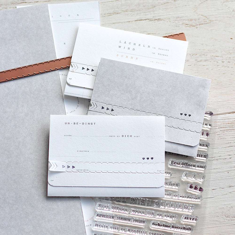 Minimalistisches Briefpapier von Alexandra Renke mit zarten Prägungen, neutralen Farbtönen und kleinen dekorativen Stempeln auf hochwertigem Designpapier, elegant präsentiert auf einer hellen Holzoberfläche.