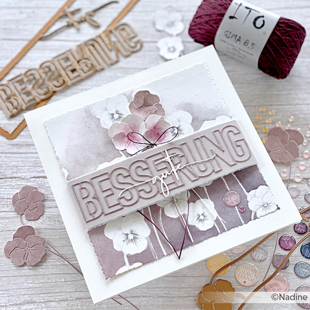 Eine handgefertigte Grußkarte von Alexandra Renke zeigt "BESSERUNG" in fetten Buchstaben, florale Stiefmütterchen-Motive und ist umgeben von Bastelmaterial.