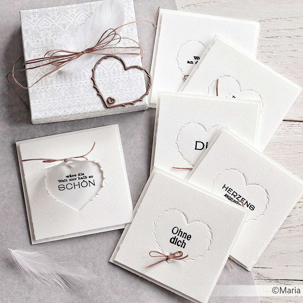Weiße Karten mit Herzausschnitten und deutschem Text, gebunden mit Schnur, sitzen neben einer Geschenkbox und weißen Federn - perfekt zum Basteln mit Alexandra Renke Bastelzubehör oder Ihrem Lieblings-Photopolymer-Stempel.