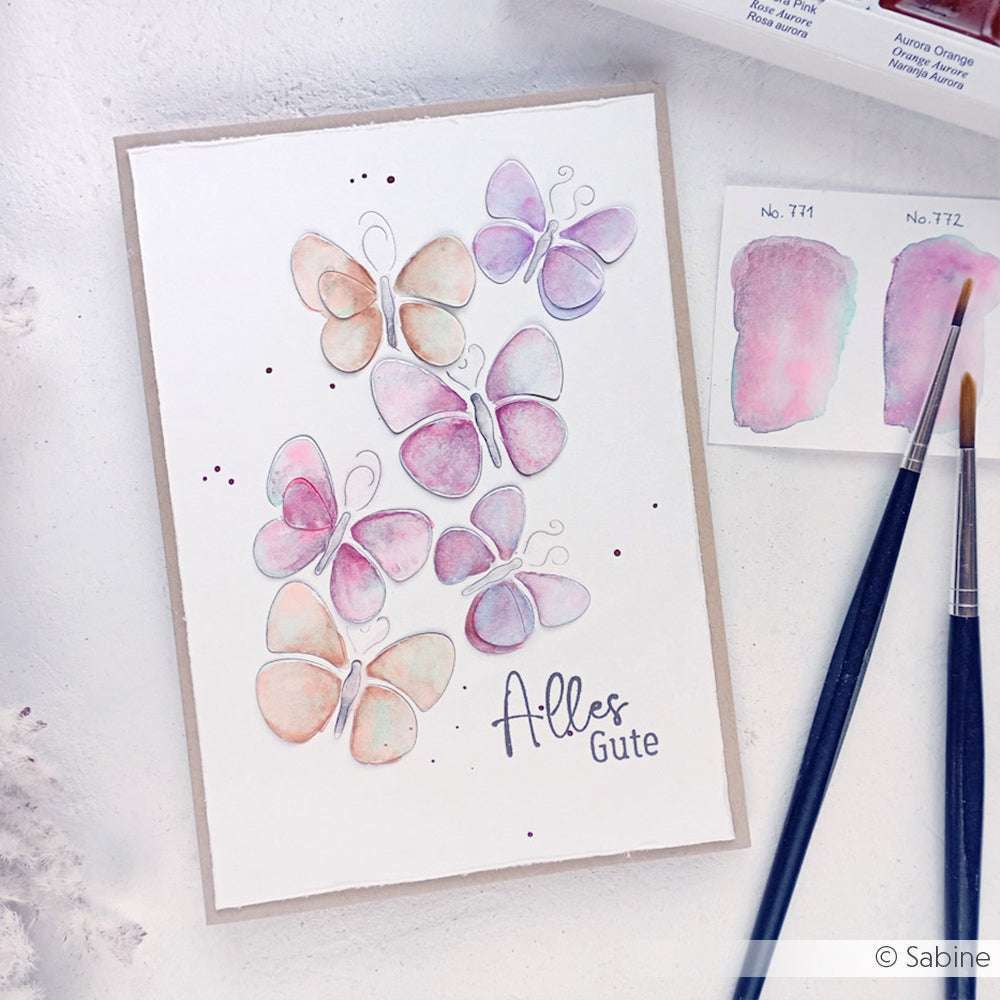 Eine Aquarellkarte von Alexandra Renke mit pastellfarbenen Schmetterlingen und "Alles Gute" auf Deutsch, umgeben von Farbklecksen, Pinseln und einem Clear Stamp für scharfe, kreative Designs.
