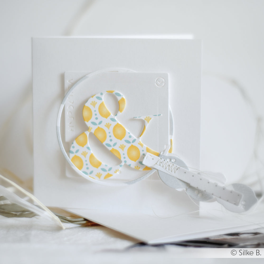 Die weiße Grußkarte von Alexandra Renke zeigt ein gelbes und blaues geblümtes kaufmännisches Und-Zeichen, das mit einer Motivstanze und einem weißen Papierschlüssel verziert ist.