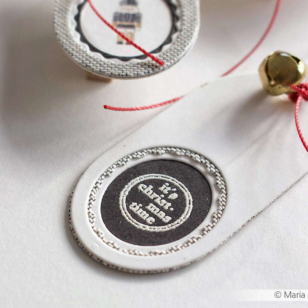 Eine Nahaufnahme von zwei handgefertigten Geschenkanhängern von Alexandra Renke, einer davon mit einer Glocke und den Worten "its charm and fun", perfekt für ein fröhliches und strahlendes Weihnachtsfest.