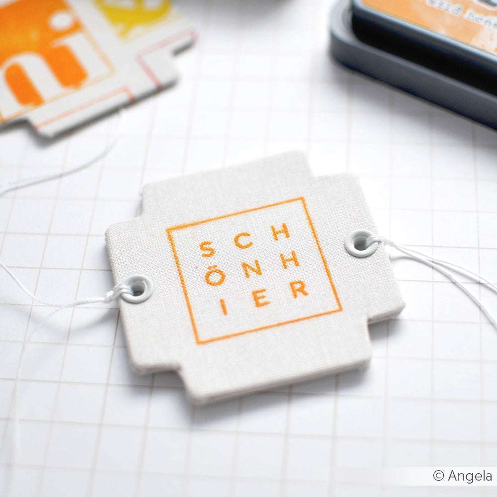 Ein Stoffetikett von Alexandra Renke mit "SCHÖNIER" in Orange, befestigt an einer weißen Schnur auf einer gerasterten Fläche - ideal für Papierprojekte oder zum Kombinieren mit Ihren Lieblingsmotiven von Clear Stamp.