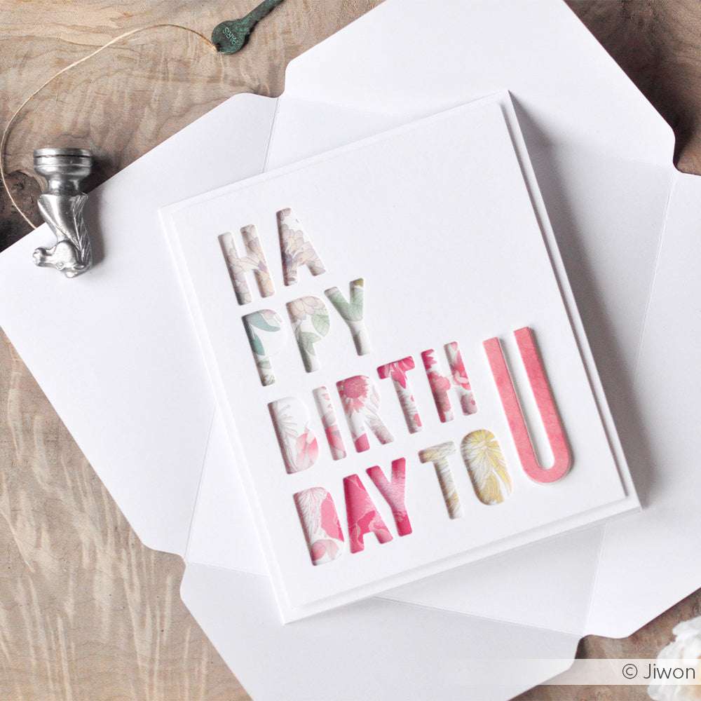 Weiße Grußkarte mit ausgeschnittenen Buchstaben "HAPPY BIRTHDAY TO U" vor einem floralen Hintergrund, gestaltet mit Alexandra Renke Designpapieren und der Motivstanze Schmales Alphabet DieCut Systeme für einen kreativen, einzigartigen Look.
