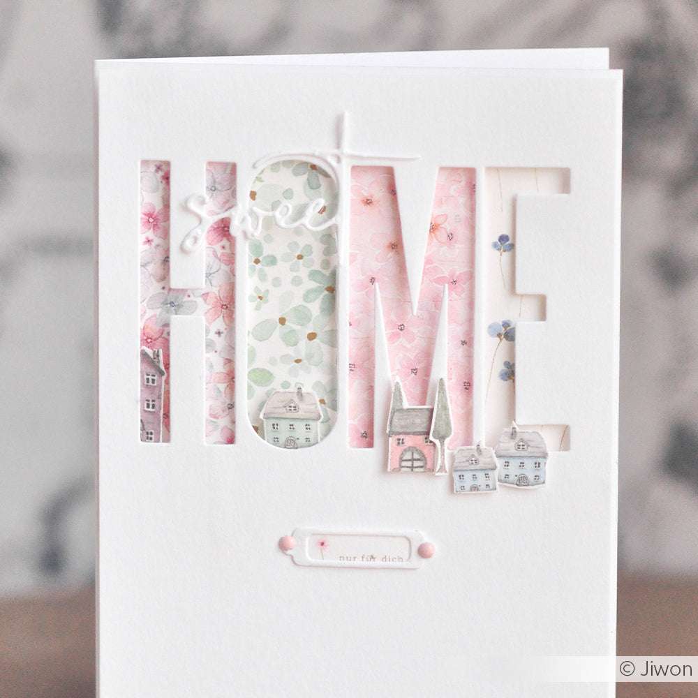 Weiße Karte mit "HOME", ausgestanzt mit der Alexandra Renke Motivstanze Schmales Alphabet, die hinter jedem Buchstaben florale Designpapiere und kleine Hausillustrationen zeigt. Ideal für kreative Stanzprojekte mit Stanzsystemen.