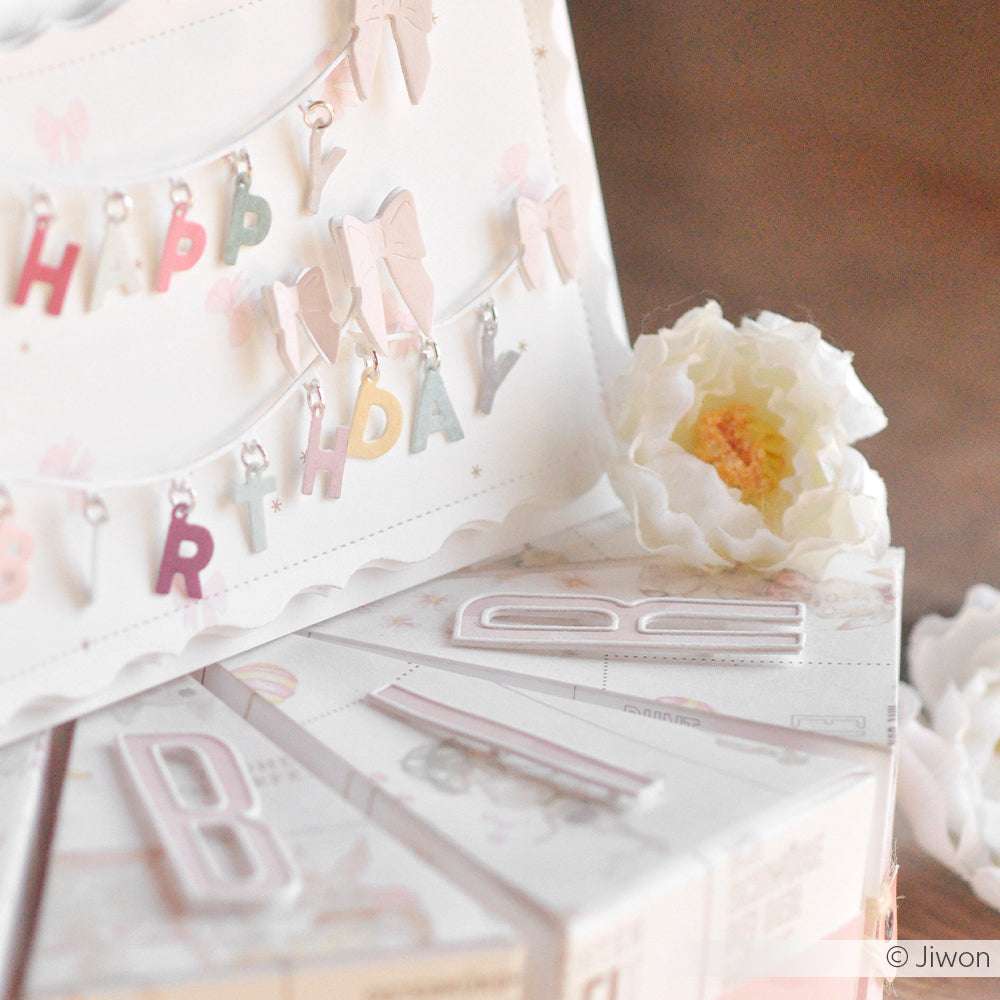 Pastellfarbenes Happy Birthday-Banner, weiße Blumen und mit Buchstaben verzierte Geschenke auf einem Tisch, gestaltet mit Alexandra Renke Designpapiere und Motivstanze Schmales Alphabet Outline DieCut Systeme für eine stilvolle Feier.