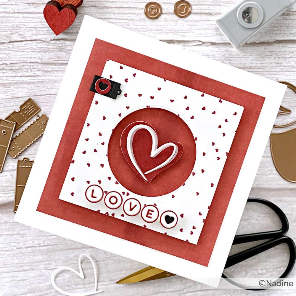 Handgemachte Valentinskarte mit Alexandra Renke Designpapier, geschichteten Herzen aus einer Herzstanze, LOVE-Buchstaben und roten, weißen und herzförmigen Mustern, die auf einem Tisch angeordnet sind.