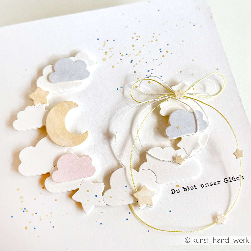 Handgemachte Alexandra Renke Karte mit Wolken, Sternen, Halbmond und einer Schleife auf weißem Hintergrund mit Farbspritzern. Der Schriftzug "It's a girl" in zarten Buchstaben wurde mit einem hochwertigen Klarsichtstempel für perfekte Details hergestellt.