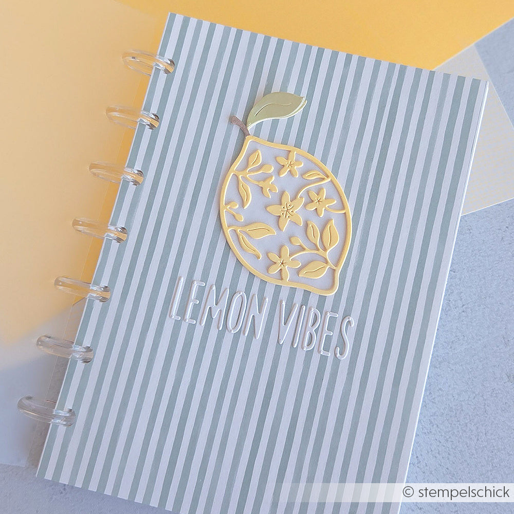 Gestreiftes Notizbuch mit einer goldenen Zitrone und dem Schriftzug „LEMON VIBES“ auf dem Einband, platziert auf einem hellen Untergrund – ideal, um es mit deinen Lieblings-Designpapieren zu kombinieren oder ihm mit der Motivstanze „Sans Serif Alphabet“ eine persönliche Note zu verleihen.