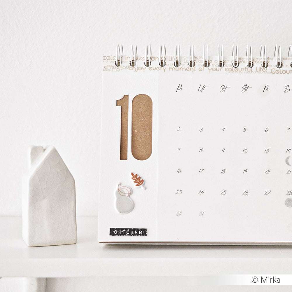 Auf einem weißen Regal stehen ein minimalistischer Oktober-Tischkalender und eine kleine weiße Hausfigur mit eleganten Zahlen, die mit Alexandra Renke Schmale Zahlen Outline Dies hergestellt wurden.
