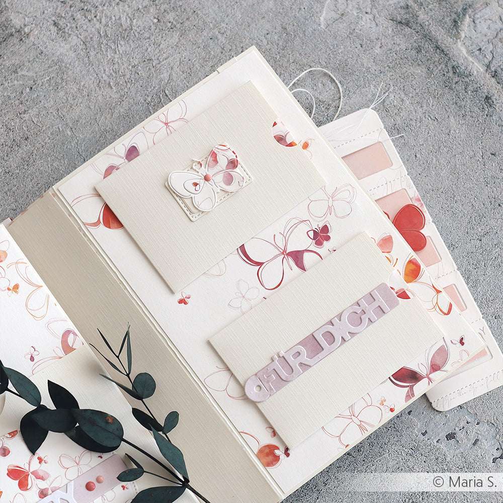 Aufgeschlagenes Scrapbook von Alexandra Renke mit Papieren im Blumendesign, Umschlägen und einem "Für Dich"-Etikett auf einer grauen, mit grünen Blättern akzentuierten Oberfläche.