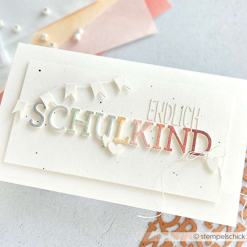 Weiße Karte von Alexandra Renke mit erhabenem Text "ENDLICH SCHULKIND" aus der Motivstanze Slab Alphabet, verziert mit pastellfarbenen Wimpeln und zwei kleinen Herzen.