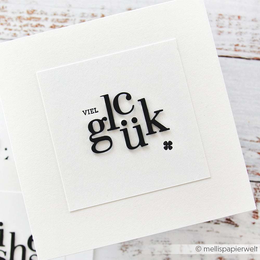 Eine weiße Karte von Alexandra Renke aus Designpapier zeigt "viel glück" in schwarzer Schrift auf einem Quadrat, das mit Motivstanze Serifbuchstaben gestochen scharf gestaltet ist.