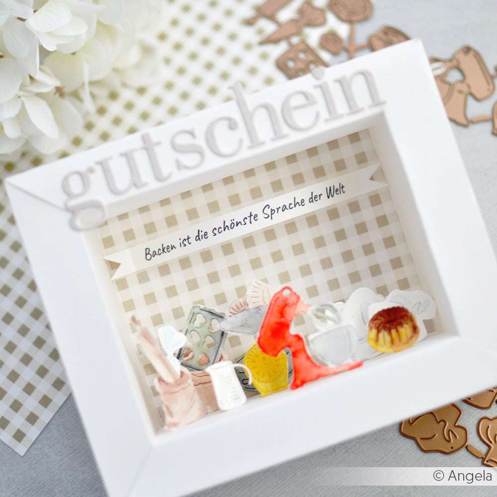 Ein weißer Schachtelrahmen von Alexandra Renke mit Backminiaturen, gestaltet mit Motivstanze Serifbuchstaben, kleinen und eleganten Designpapieren, beschriftet mit "gutschein" und "Backen ist die schönste Sprache der Welt".