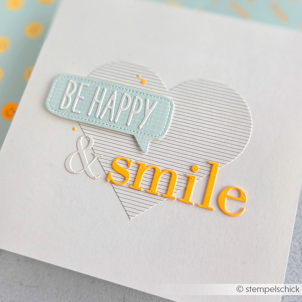 Weiße Karte aus hochwertigem Designpapier mit einem gestreiften Herz, der Sprechblase „BE HAPPY“ sowie fröhlichen orangefarbenen Buchstaben und einem Lächeln. Perfekt abgerundet durch die Motivstanze „Serifbuchstaben“ für einen charmanten Touch – ideal für kreative Stanzsysteme-Projekte.