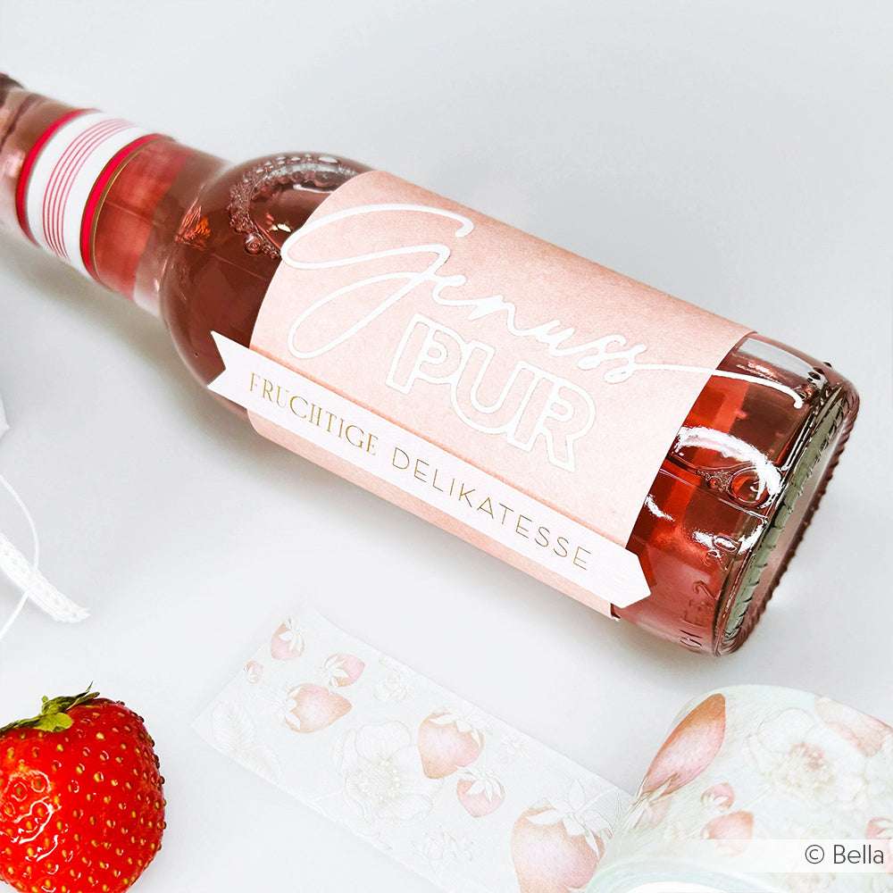 Ein rosafarbener Flakon mit der Aufschrift Genuss Pur von Alexandra Renke steht neben einer Erdbeere und einer Schleife mit Fruchtmuster, die mit eleganten Designpapieren veredelt wurde und für zusätzlichen Stil sorgt.